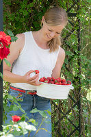 Woman with fresh strawberries in bowl under rose arch in summer garden