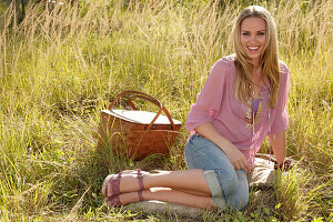 Blonde Frau in rosa Bluse und Jeans-Caprihose mit Picknickkorb auf der Wiese