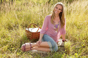 Blonde woman in pink blouse and jeans capri pants with picnic basket on the meadow