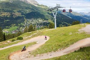 Mountainbiker, die eine Mountainbike-Strecke an der Station Lenzerheide hinunterfahren, mit den Seilbahnen und den Schweizer Alpen im Hintergrund, Lenzerheide, Kanton Graubünden, Schweiz