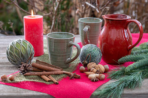Weihnachtlichte Tischdeko mit Kaffeetassen, Zimtstangen, Christbaumkugeln, Nüssen, Tannenzweig und Kerze