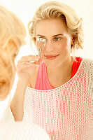 Blonde woman wearing a pink top and white summer sweater uses an eyelash curler