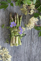 Bund grüner Spargel mit Storchenschnabel (Geranium) und Holunderblüten (Sambucus Nigra) auf Holzuntergrund