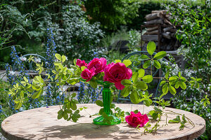 Rote Rosenblüten (Rosa) dekoriert mit Stein auf Stielvase und Zweigen auf Gartentisch