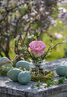 Rosenblüte (Rosa) mit Zweigen in Vase und Ostereiern auf Gartentisch