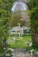 Gedeckter Tisch im Garten für Osterfrühstück mit Osternest und gefärbten Eiern, Blumenstrauß, Hund in der Wiese, Torbogen durch Kletterpflanzen