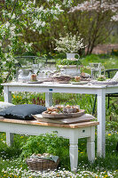 Set table in the garden for Easter breakfast with Easter nest and coloured eggs in egg cups and bouquet of flowers in etagere, basket with eggs and parsley in meadow