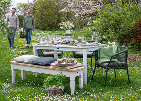 Young couple in front of a laid table, Easter breakfast with Easter nest and coloured eggs in egg cups and parsley in a basket in the garden