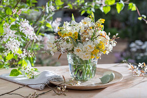 Blumenstrauß aus Schlüsselblume (Primula veris), Felsenbirne (Amelanchier), Narzissen 'Bridal Crown' (Narcissus) in Glasvase und Eierschale auf Gartentisch