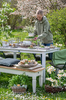 Junge Frau stellt Blumenstrauß auf gedeckten Tisch zum Osterfrühstück mit Osternest, Weidenkorb mit Petersilie und gefärbten Ostereiern und Narzissen in Blumenwiese
