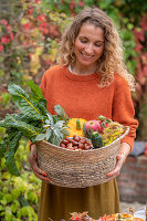 Frau hält Korb mit Zucchini, Kürbis, Äpfeln, Kastanien, Fenchelblüten und Mangold