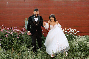 Bride and groom holding hands against brick wall and flowers