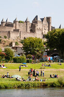 Menschen versammeln sich auf der Wiese am Fluss mit dem Schloss und den Festungsmauern im Hintergrund; Carcassonne, Languedoc-Rousillion, Frankreich