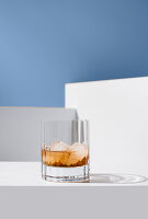 Nahaufnahme eines transparenten Whiskeyglases mit Eiswürfeln auf einer weißen Fläche vor weißen Wänden