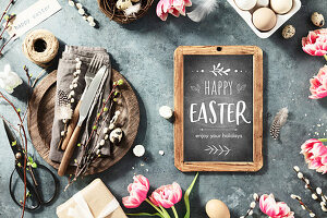 Easter table with Happy Easter text on chalkboard, spring flowers and cutlery on dark blue background top view flat lay