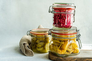 Glass jars of fermented cabbage with beetroot, spicy peppers, and white cucumbers on a wooden board.