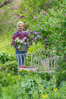 Frau mit Blumenstrauss aus Fliederblüten (Syringa) vor Gartenbank