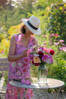 Frau an Gartentisch und bunter Blumenstrauß aus Dahlien (Dahlia), Rosen (Rosa), Herbstanemonen (Anemone Hupehensis) und wilde Möhre