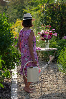 Frau mit Gießkanne vor Gartentisch mit Blumenstrauß aus Dahlien (Dahlia), Rosen (Rosa), Herbstanemonen (Anemone Hupehensis) und wilde Möhre