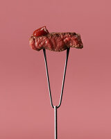 Peace of appetizing rare steak with ketchup on stainless steel meat carving fork against pink background