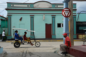 Kuba,Baracoa,ein Mann sitzt an der Straße und beobachtet ein Dreiradtaxi, das vor einem bunten Gebäude vorbeifährt