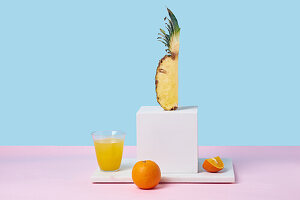 Quarter of a pineapple on a white pedestal surrounded by oranges and orange juice