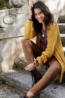 Langhaarige junge Frau in maisgelber Strickjacke, brauner Hose und Buntem Shirt