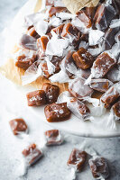 A close-up shot of a large pile of homemade caramel candies on a marble surface. The candies are wrapped in white parchment paper and sprinkled with sea salt. Arranged on a piece of brown parchment paper, which is placed on a white marble cake stand. The blurry background shows more caramel candies, suggesting a delicious treat spread and the simplicity and beauty of homemade sweets.