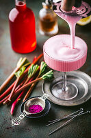 Action shot of frothy pink rhubarb cocktail being poured from shaker into beautiful full coupe glass with fresh rhubarb and red syrup in background