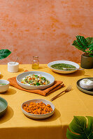 A sumptuous Italian dinner with bowls of basil pesto gnocchi, fresh green salad, chunks of sweet potato and creamy ricotta cheese. The scene is set on a bright yellow tablecloth against an orange background with green leaves.