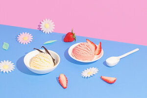 Two bowls of vanilla and strawberry ice cream against a bright pink and blue background, garnished with fresh strawberries, decorative flowers and a white spoon.