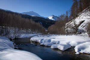 Winterliche Berglandschaft mit Bach und schneebedeckten Gipfeln in Val Müstair (Schweiz)