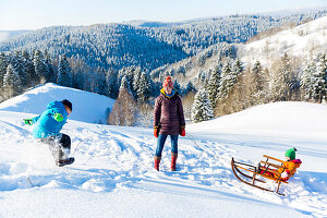 Young family in the snow in winter, boy jumping in the snow, mother pulling daughter on sledge, Harz, MR, Sankt Andreasberg, Lower Saxony, Germany