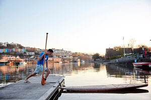 Mann mit Paddel in der Hand macht Dehnübung am Flussufer, bevor er auf das Paddleboard steigt