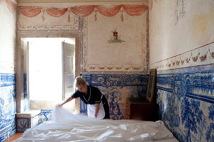 A maid is making the bed in a frescoed room, with light streaming from an open window. Shot inside Palacio Belmont, Lisbon, Portugal.