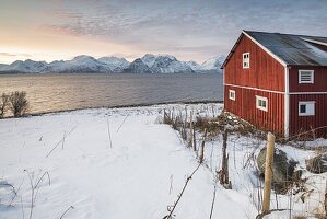 Typisches Rorbu in der verschneiten Landschaft bei Sonnenuntergang auf dem Fjord, Djupvik, Lyngen Alpen, Tromso, Norwegen, Europa