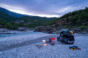 Albania, Southern Europe, young couple sitting in front of off-road vehicle with roof tent, campfire, river, Vjosa, Permet