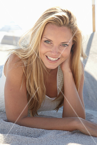 A Mature Blonde Woman Lying On A Sandy Beach Wearing A White Top